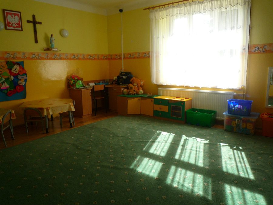 Przestronna sala z zieloną wykładziną, krzyżem i godłem na ścianie. Przez duże okno wpada słońce.