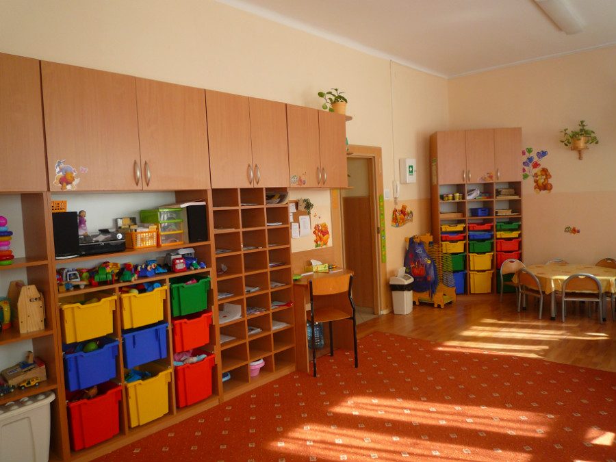 Jasna sala z regałami z kolorowymi pojemnikami na zabawki i stolikami przy oknie.