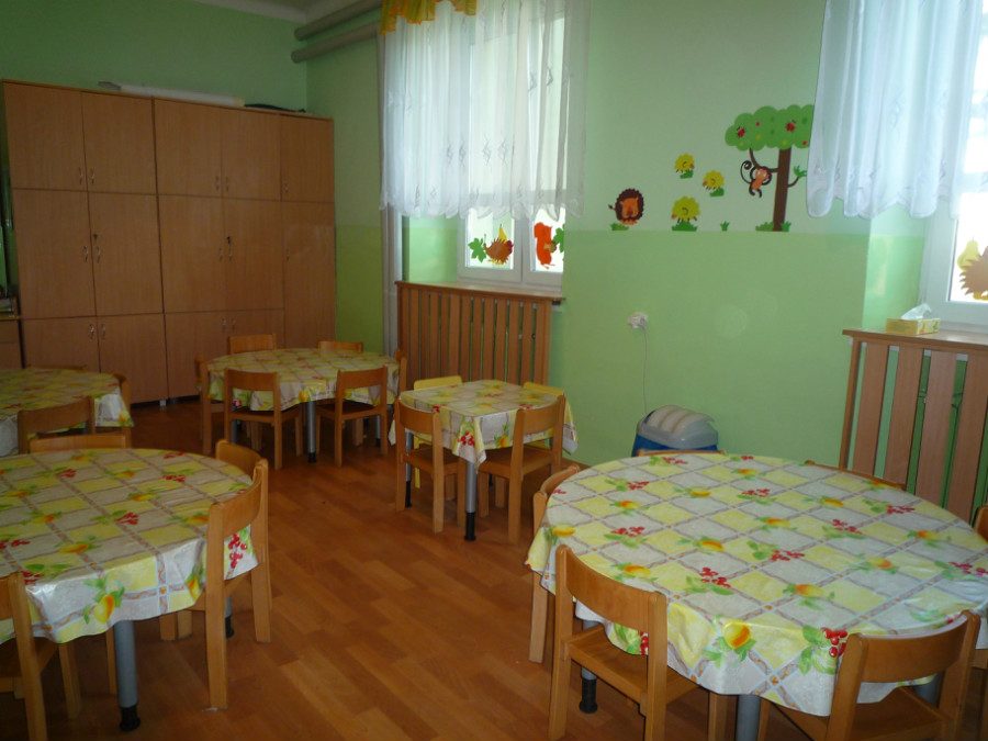 Sala z zielonymi ścianami, okrągłymi stolikami nakrytymi obrusami i drewnianymi krzesełkami dla dzieci.